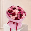 A large bouquet of pink and red flowers wrapped in light pink paper and tied with a dark red ribbon, displayed on a white pedestal against a soft beige background.