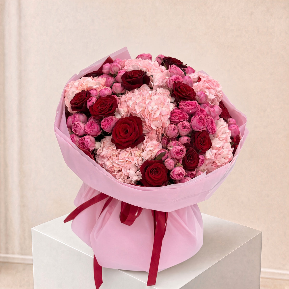 A large bouquet of pink and red flowers wrapped in light pink paper and tied with a dark red ribbon, displayed on a white pedestal against a soft beige background.