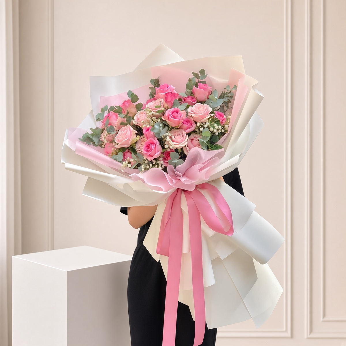 A large bouquet of pink roses wrapped in white and pink paper with a pink ribbon, held by a person wearing a black dress in front of a beige paneled wall.