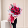 A large bouquet of red roses wrapped in pink and white tissue paper with a pink ribbon, held by a person standing behind a white pedestal against a beige paneled wall.