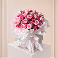 A large bouquet of pink and white roses wrapped in white and pink tissue paper, tied with a white ribbon, displayed on a white pedestal against a soft beige paneled background.