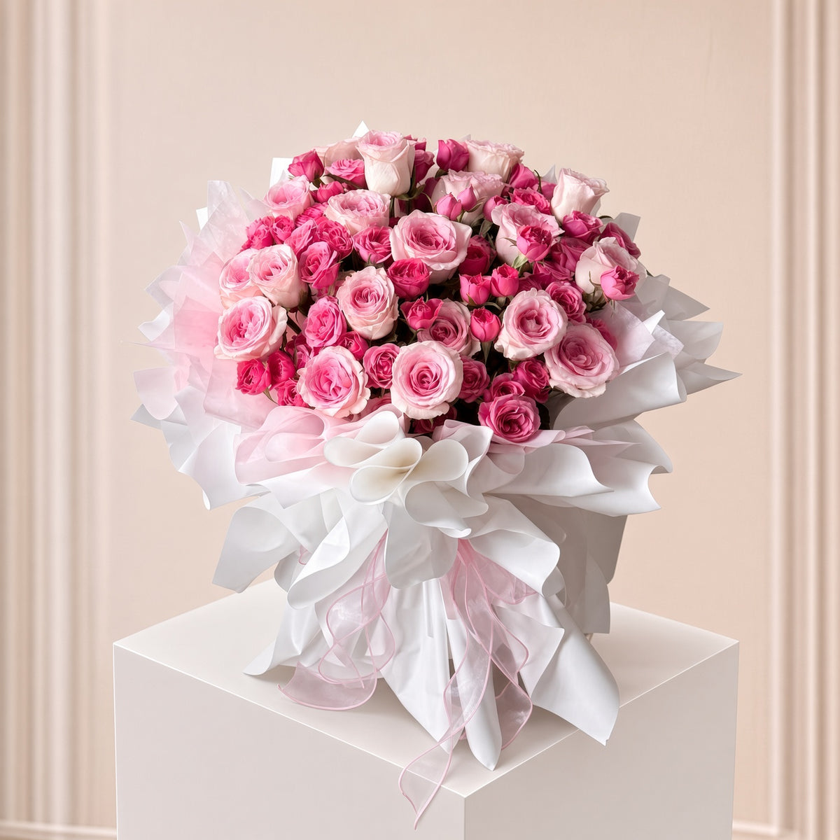 A large bouquet of pink and white roses wrapped in white and pink tissue paper, tied with a white ribbon, displayed on a white pedestal against a soft beige paneled background.