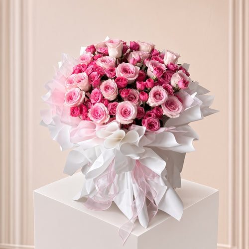 A large bouquet of pink and white roses wrapped in white and pink tissue paper, tied with a white ribbon, displayed on a white pedestal against a soft beige paneled background.