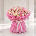 A large, vibrant floral arrangement of pink, peach, and cream roses wrapped in layered pink paper and tied with a white ribbon, set against a bright, elegant white interior background.
